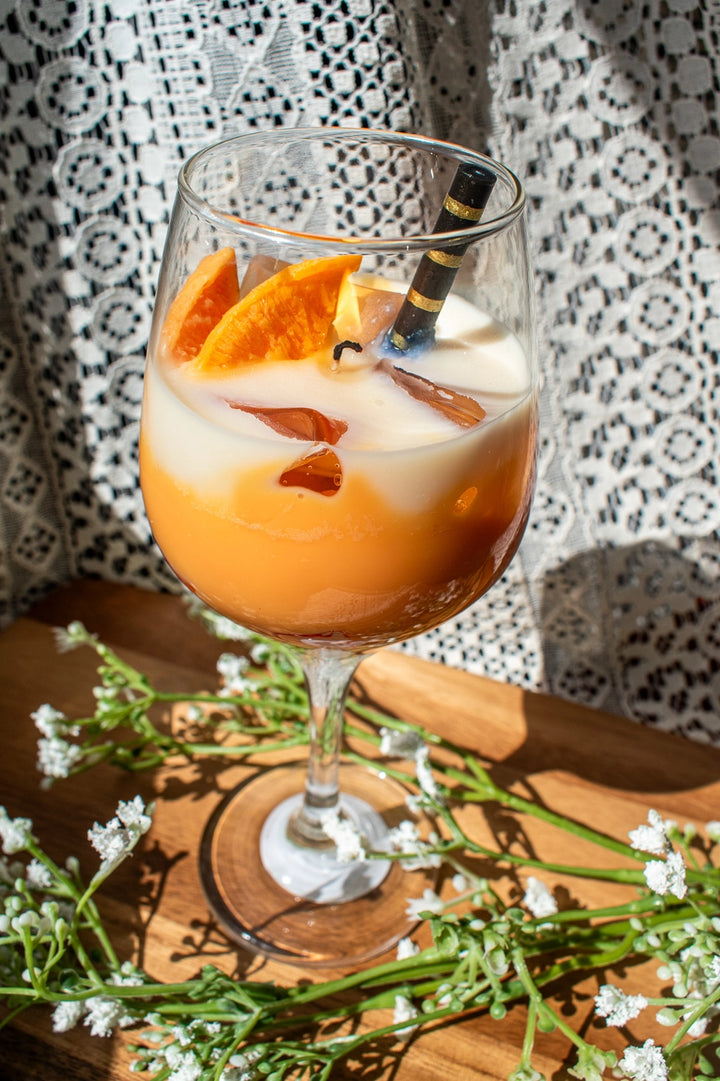 aeperol spritz inspired candle. Glass of orange cocktail with ice and garnish on a wooden board with flowers.